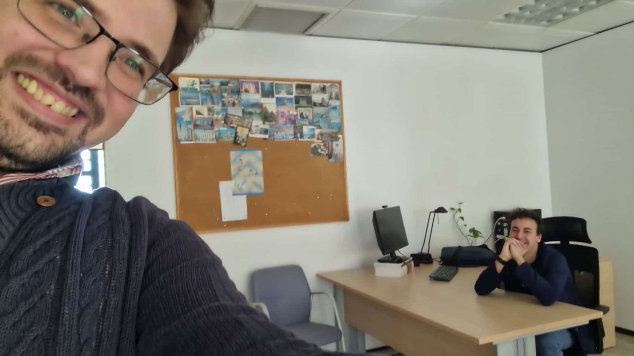 Selfi en un despacho universitario con un compañero sentado al fondo